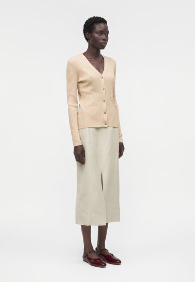 Γυναίκα που στέκεται, φοράει μπεζ ριμπ δαντελωτή ζακέτα με κουμπιά, κρεμ midi φούστα με άνοιγμα μπροστά και μπορντό Mary Jane flats σε απλό φόντο.