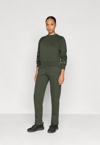 Olívazöld egyforma pulcsi és nadrág szett puha anyagból, bordázott mandzsettákkal és minimalista logóval a mellkason és a lábon.