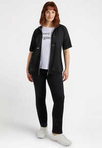 Black zip-up jacket with short sleeves, featuring shiny accents. Worn over a white t-shirt with black text. Paired with dark pants and white sneakers.