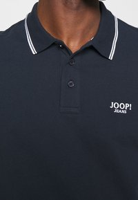 Marinblå polo t-shirt i strukturerad tyg, med en spetsig krage med vita ränder och en liten vit logotyp på vänster sida.