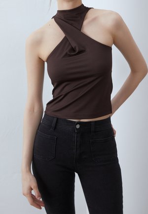 Woman wearing a sleeveless dark brown asymmetrical halter top paired with high-waisted black jeans against a plain background.