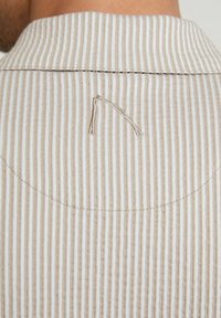 Camisa de tela a rayas con una combinación de colores beige y blanco, que presenta un sutil acento bordado en el cuello trasero.