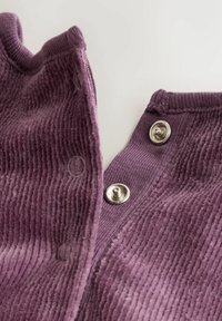 Közelről készült felvétel lila kordbársony anyagról, két ezüst snap gombbal, amelyen láthatóak a gomblyukak és a texturált bordás minta egy világos háttéren.