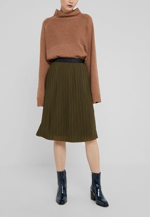 Vrouw draagt een bruine trui die in een olijfgroene geplooide rok is gestopt en marineblauwe enkellaarsjes van lakleer, staand tegen een effen grijze achtergrond.