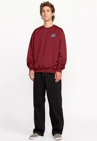 Sweat-shirt bordeaux avec poignets et ourlet côtelés, arborant un petit patch logo sur la poitrine. Assorti à un pantalon noir ample et des chaussures à carreaux.