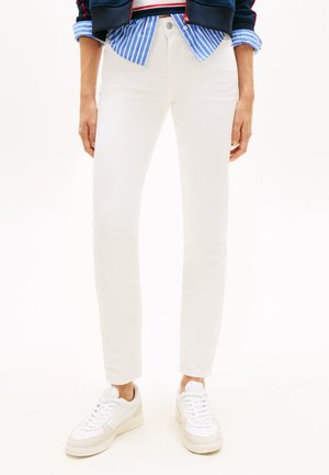 Osoba v bílých kalhotách se širokými nohavicemi, modře pruhované košili a bílých teniskách stojící před jednobarevným pozadím.