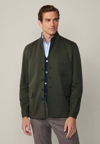 Giacca verde scuro con colletto alla cinese, bottoni frontali e due tasche laterali. Indossata sopra una camicia blu e un gilet blu navy.