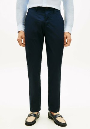 Uomo che indossa pantaloni eleganti blu navy, camicia a righe azzurro chiaro infilata e mocassini beige e neri, in piedi davanti a uno sfondo neutro.