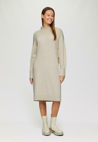 Beige gebreide jurk met lange mouwen, hoge hals en een rechte silhouette, gecombineerd met gladde, lichtgekleurde enkellaarsjes.