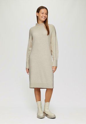 Beiges Strickkleid mit langen Ärmeln, hohem Kragen und gerader Silhouette, kombiniert mit glatten, hellen Ankle Boots.
