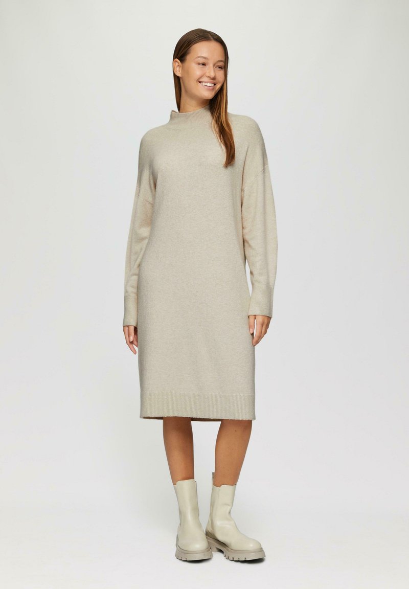 Beige gebreide jurk met lange mouwen, hoge hals en een rechte silhouette, gecombineerd met gladde, lichtgekleurde enkellaarsjes.