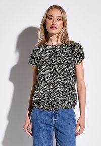 Schwarz-creme gemusterte Kurzarmbluse mit rundem Ausschnitt und Raffung am Saum, kombiniert mit blauen Jeans.