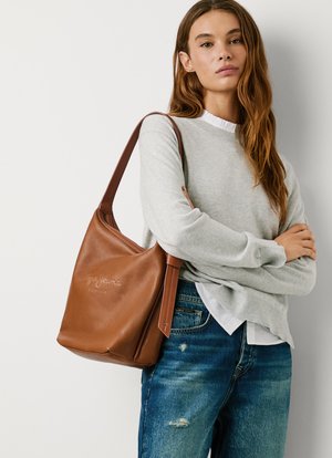 Borsa a spalla in pelle marrone con una forma morbida, dotata di una parte superiore piatta, una tracolla singola e dettagli del logo impresso. Indossata con un maglione grigio chiaro e denim.