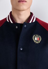 Marineblå og bordeaux varsity-jakke med stribet krave, trykknapper og broderet logo "TOMMY NEW YORK" med dekorativ laurbærkrans.