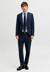 Tumesinine ülikond, kitsas lõige, ühe rinnaga jakk ja triibuline särk. Mustad nahast kingad paeladetailidega.