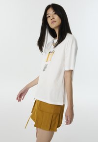 T-shirt en coton blanc avec un design graphique représentant des crayons de couleur, associé à un short jaune moutarde avec poches et une finition texturée.