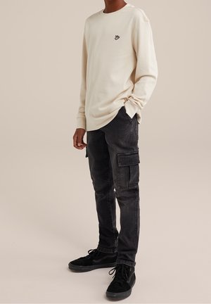 Person trägt ein beigefarbenes Langarmshirt mit einem kleinen Hand-Logo, schwarze Cargohosen und schwarze Turnschuhe und steht vor einem schlichten Hintergrund.