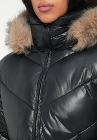 Nahaufnahme einer Person, die eine schwarze Daunenjacke mit einem Reißverschluss vorne und einer Kapuze mit Fellbesatz trägt.