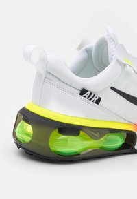 Bílé tenisky s černým Swoosh, průhledným zeleným polstrováním a jasně žlutou podešví. Texturovaný textilní svršek s poutkem.