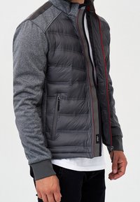 Mann trägt grau gesteppte Jacke mit gestrickten Ärmeln, Reißverschlusstasche und vorne mit rotem Rand versehenen Reißverschluss, kombiniert mit weißem Hemd und schwarzer Hose.