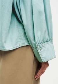 Gros plan sur une personne portant une chemise à manches retroussées bleu clair et un pantalon beige, la main détendue le long du corps.