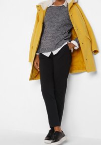 Personne portant un manteau jaune par-dessus un pull gris, une chemise blanche, un pantalon noir et des baskets noires, debout avec une main dans la poche contre un mur blanc.