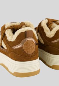 Sneaker in suede marrone con una soffice fodera beige, lacci rotondi e un logo in rilievo sul tallone. Presentano una suola stratificata bianca e beige.