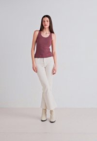 Top in tessuto bordeaux con bordi scallop, abbinato a pantaloni bianchi a campana e stivali beige alla caviglia, in piedi contro uno sfondo chiaro.