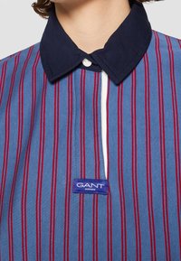 Polo rayé verticalement en bleu et rouge, avec un col marine et une patte de boutonnage blanche ; étiqueté avec un label GANT bleu en bas.