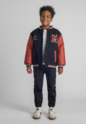 Veste varsity noire et rouge avec des manches en cuir, col côtelé blanc et lettre 'M' brodée, associée à un pantalon cargo et des baskets.