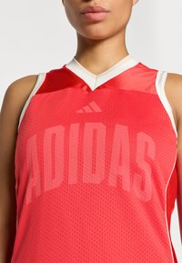 Κόκκινη αθλητική φανέλα με πλέγμα, σχεδιασμένη με λευκή επένδυση και μεγάλο, τονικό λογότυπο «ADIDAS» στο μπροστινό μέ ρος.