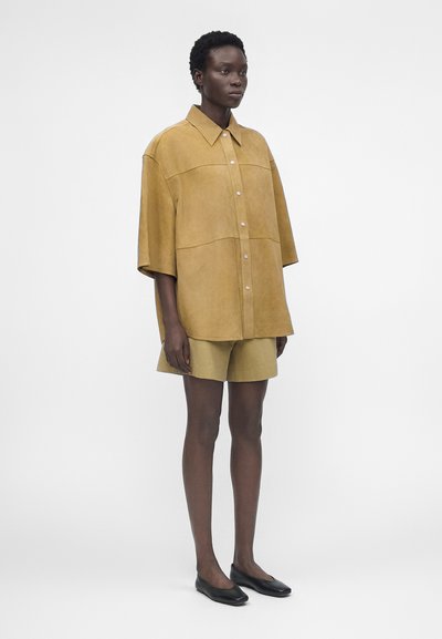 Άτομο με σκουρόχρωμο δέρμα φοράει υπερμεγέθη ταμπά πουκάμισο με κουμπιά, ταιριαστά σορτς και μαύρα φλατ παπούτσια, στέκεται μπροστά σε απλό λευκό φόντο.