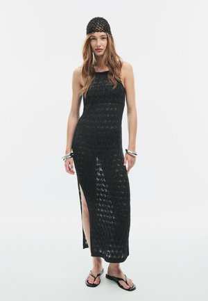 Femme portant une robe noire sans manches en crochet avec une fente latérale, une casquette noire en crochet, des bracelets argentés et des tongs noires, debout sur un fond blanc.