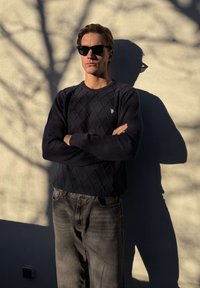 Junger Mann mit Sonnenbrille, dunklem Pullover und Jeans steht mit verschränkten Armen vor einer sonnenbeschienenen Wand, die Baumschatten wirft.