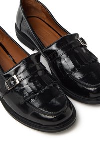 Mocasines de charol negro con un acento de hebilla y diseño con flecos, con una textura suave y suela de goma de perfil bajo.