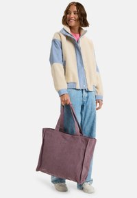 Torba tote z prążkowanej fioletowej tkaniny, z dwiema bocznymi rączkami i prostym akcentem logo. Nosi ją osoba w beżowej i niebieskiej kurtce oraz dżinsach.