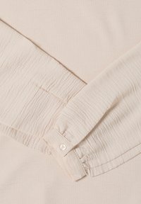 Jasnobeżowa, teksturowana tkanina o pomarszczonym wyglądzie. Widoczny mankiet z guzikiem oraz złożony rękaw. Delikatny wzór dodaje głębi.