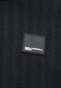 Melna adīta auduma virsma satur paceltas vertikālas svītras un taisnstūra melnu etiķeti ar uzrakstu "KARL LAGERFELD JEANS" baltā krāsā.