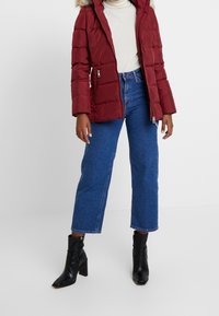 Röd vadderad jacka med huva och fuskpälsdetalj, kombinerad med midblå caprijeans och svarta ankelboots, med en blank yta.