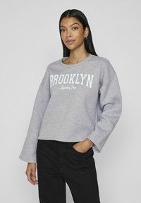 Suéter gris claro con la palabra "BROOKLYN" impresa en blanco a lo largo del pecho. Presenta mangas largas y sueltas y un cuello redondo, combinado con pantalones negros.
