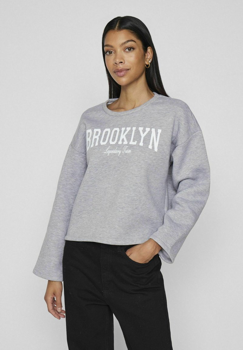 Suéter gris claro con la palabra "BROOKLYN" impresa en blanco a lo largo del pecho. Presenta mangas largas y sueltas y un cuello redondo, combinado con pantalones negros.