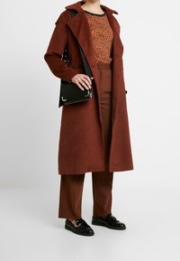 Dlouhý kabát z vlny v rezavé barvě s límcem, v kombinaci s oranžovým vzorovaným topem a hnědými kalhotami. Černá taška a boty dokončují outfit.