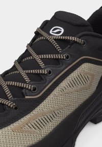 Chaussure de sport noire et beige avec des lacets à motifs, un panneau latéral en mesh, une languette rembourrée et un logo sur le dessus avant.