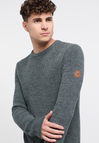 Maglione grigio lavorato a maglia con scollatura rotonda e polsini a coste. Presenta una piccola etichetta con logo marrone sulla manica sinistra. Tessuto morbido.