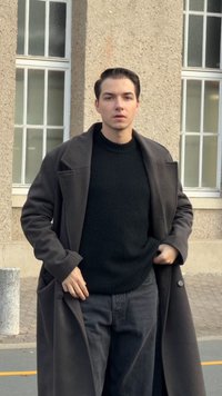 Tumša vilnas mētelis pār melnu adītu džemperi, pelēkas džinsa bikses, stāvot priekšā akmens sienai. Mētelim ir plata apkakle un pogu detalizējums.