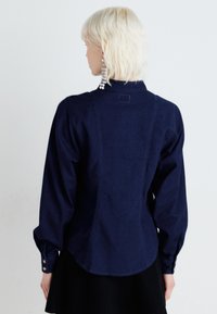 Camicia in denim blu scuro con colletto alto, maniche lunghe a sbuffo e chiusura con bottoni. Presenta una cucitura decorativa lungo la schiena. Texture liscia.