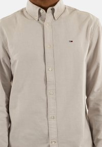 Camicia beige con bottoni in cotone, dotata di colletto classico, con bottoni bianchi e un piccolo logo ricamato in rosso, bianco e blu.