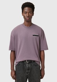 Camiseta oversized de color lavanda hecha de algodón. Mangas cortas, cuello redondo, con un detalle del logo negro en el pecho. Combinada con jeans oscuros.