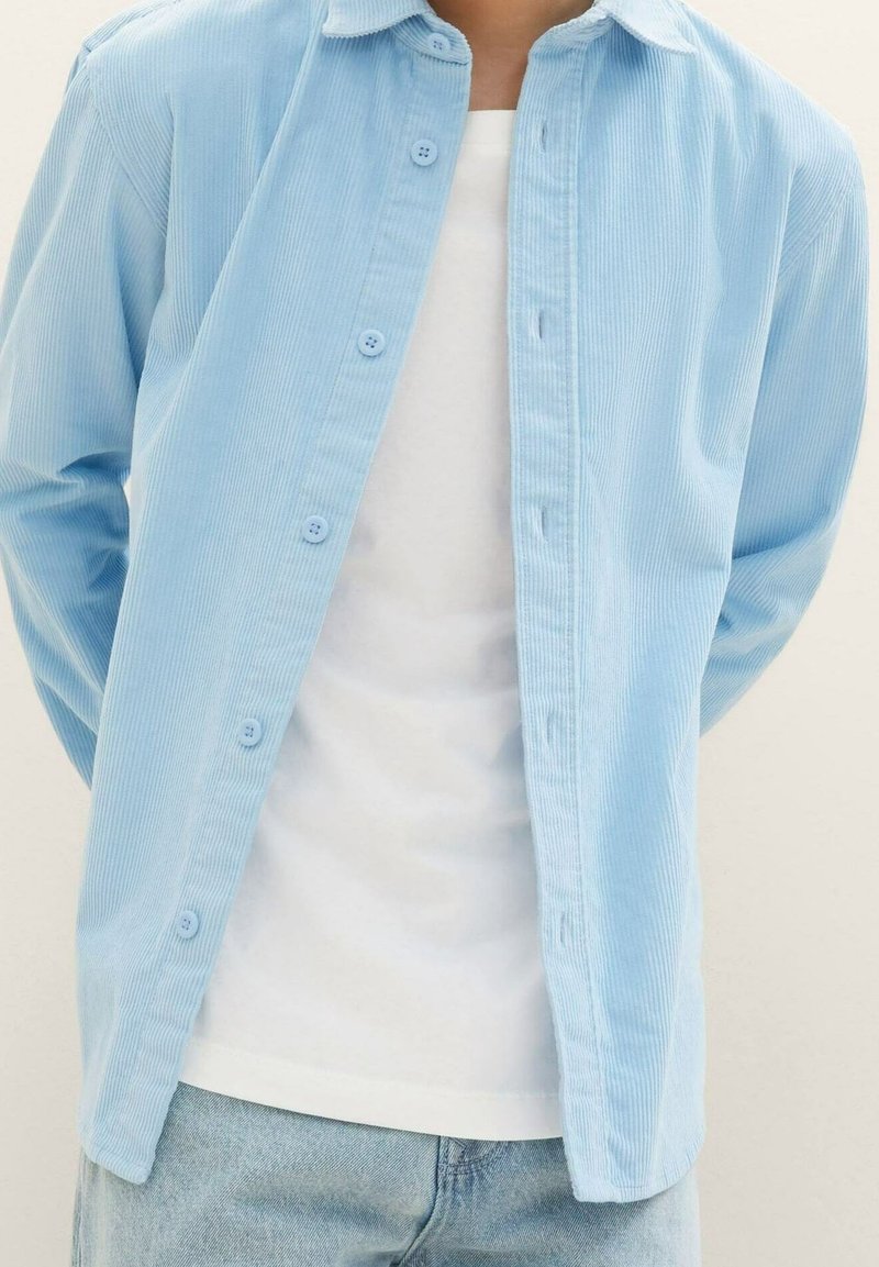 Personne portant une chemise en velours côtelé bleu clair boutonnée sur un t-shirt blanc et un jean bleu clair, mains dans le dos, fond neutre.