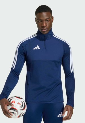 Mand i mørkeblå Adidas træningsdragt holder en hvid og sort Adidas Tiro fodbold mod en ensfarvet baggrund.